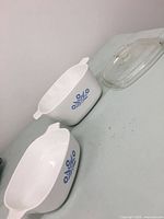 Two white ceramic CorningWare Blue Cornflower dishes with blue floral pattern, one larger one smaller, and a clear glass lid, all arranged on a flat surface.