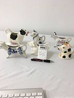 Front view of six mixed color ceramic cow creamers arranged in two rows, showing details and scale via pen