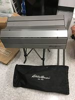 Top angled view of folded aluminum slat table on legs with black Eddie Bauer branded carrying bag below.