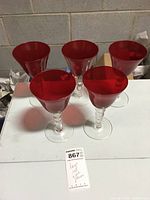 Five red glass goblets with clear beaded stems arranged on a white surface.
