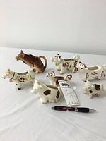 Seven vintage cow creamers of various sizes and painted designs arranged on a white surface with a pen for size comparison. All are Little Brown Cow style creamers with handles formed as tails and creamer spouts at the cows' mouths.