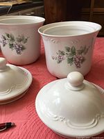 Photo showing two Pfaltzgraff canisters with lids, grape design, white ceramic, placed on a red cloth background.