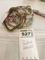 Bracelet lot displayed on a table with a handwritten lot tag reading 'Pinkish Bracelets'. Various bracelet styles visible including ceramic floral, faux pearl, flower bead, and gold-tone bangles.