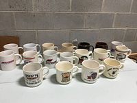 Photo showing all 18 ceramic mugs arranged in rows on a white surface against a gray block wall background, displaying various logos and colors ranging from white, cream, black, and maroon mugs.