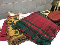 Photo showing folded red, green, and yellow-green floral woven fabric placemats with fringed edges on top of a pile of placemats and items.