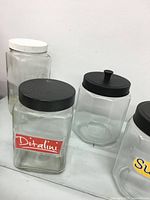 Set of six clear glass jars in various shapes and lid types, including plastic black, white, and metal lids, with labels visible on some jars.