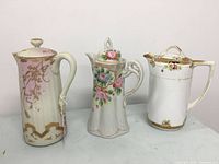 Front view of 3 Victorian-style porcelain pitchers and teapot, showing floral and gold-accented designs and mixed hand-painted patterns.