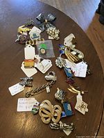 Overview of an assortment of 32 vintage style metal and enamel pins and brooches laid out on a wooden surface. Various designs include floral, geometric, bow, ship, and butterfly.