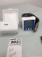 WrisTech wrist blood pressure monitor with digital display and gray strap, alongside white plastic storage case, and a pen on the side for scale.