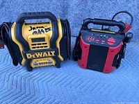 Front view of two battery boosters, one yellow and black DeWalt and one red and black unit, showing handles, displays, and cables.