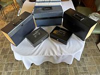 Three file boxes and two small Sentry Safe cash boxes on a white tablecloth. The file boxes are black and blue-grey with handles and locks. The cash boxes are black with Sentry Safe branding, metal, with small key locks but keys are missing.
