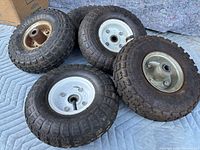 Five off-road tires with four mounted on metal rims, placed on ground for display. Tires have knobby tread, some dirt visible on rubber and rims.