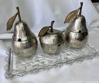 Three metallic serving dishes shaped like an apple and two pears with lids and leaf detailing, placed on a rectangular crystal tray