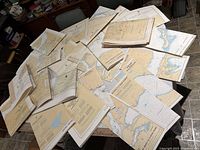 Photo showing a broad arrangement of multiple marine navigational charts spread out on a table, demonstrating their paper material and map content.