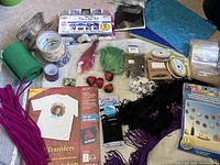 Overview of assorted craft supplies on a light surface including yarn, pipe cleaners, tie dye kit, transfers, ribbons, fake strawberries, googly eyes, and candles.