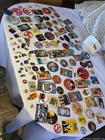 Overview of large collection of assorted vintage pins and buttons spread on a white cloth on bed, featuring multiple circular, rectangular and irregular shaped pins of various sizes and colors.