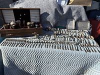 Wide top view showing multiple rows of assorted silverware laid out on a quilted pad, including spoons, forks, knives, and small silver containers in a wooden holder.