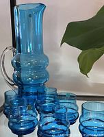 Blue blown glass pitcher and seven matching tumblers on a glass surface, some tumblers with original stickers.