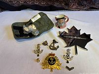 Photo showing the full lot with various hat badges, pins, a stainless steel cut Maple Leaf with Navy Destroyer cutout, a ceramic seaman mug, a pen on a metal maple leaf base, and a military sew kit pouch with camouflage paint tube.