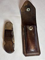Gerber folding cutter knife beside its leather sheath case, showing the handle materials and blade folded into handle