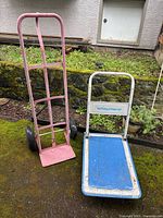 Front view of pink hand truck and blue deck platform dolly on mossy ground near stone wall