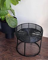 Full view of black wire patio/outdoor tray table next to a plant, showing circular woven tray top and metal legs.