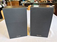 Pair of black wood grain bookshelf speakers viewed from front on white tablecloth with wood furniture background.