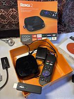 Roku 2 streaming player, remote, and adapter in orange box along with Roku streaming stick on table
