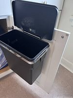 Open black garbage bin mounted inside cupboard with steel frame visible, showing side attachment.