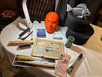 Overview of the lot including an orange mannequin head, wooden train whistles, model train car in plastic case, vintage ashtray, Carlsberg lighter, decorative fish sculpture, scrapbook of ephemera and other items, arranged on a table.