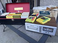 Full lot view showing 8 track deck, carrying case, and multiple 8-track tapes arranged on black table outdoors.
