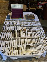 Full layout of 92-piece South Seas silverware set and additional silver utensils placed on a white tablecloth, including a fabric-covered case for Mother of Pearl cutters and a decorative tray.