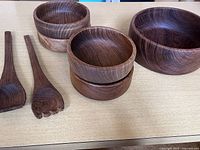 Photo showing four small teak bowls, one large teak bowl, and two wooden salad utensils (a spoon and a fork) laid out on a surface, highlighting the natural wood grain and polished finish.