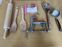 All items in the lot shown on a table, including wooden rolling pin, wooden spoon, spatula, wooden ladle, ceramic ladle, and apple peeler/corer/slicer.