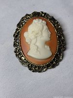 Front view of vintage cameo brooch with white profile on orange background in ornate metal frame.