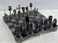 Angled side view of chess pieces on board showing silver and black automotive part chessmen, and the metal board with steel edges and rounded corner supports.