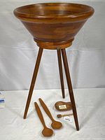 Large wooden salad bowl on a detachable three-legged stand with two wooden spoons, a honey dipper, and small acacia dish on white background.