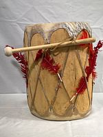 Full view of ceremonial wooden drum with red feathers attached and wooden mallet positioned horizontally on the side.