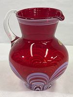 Front angle of ruby colored crystal pitcher showing clear handle, flared spout, ruby red body, and white horizontal stripes near rim.