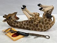 Photo showing ceramic giraffe wine bottle holder lying on its back, corkscrew, and church key bottle opener arranged together