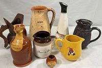 Wide view of the full collection of seven pitchers including six ceramic and one wood showing different shapes, colors, and decorative details.