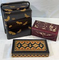 Front view of all three storage boxes displaying the Asian motif plastic box, Mexican tooled leather box, and Russian inlaid wood box.