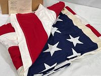 American flag folded with visible red and white stripes and blue field with white stitched stars, flag partially unpacked from original Veterans Administration cardboard box with shipping label visible.