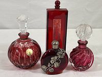 Four ruby red glass perfume bottles arranged on white surface: one round with cut glass and labeled 24% PbO hand-cut crystal, one round with silver floral decoration, one swirl cut, and one rectangular Shiseido bottle.