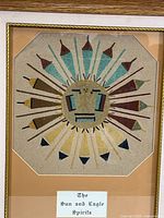 Close-up photo showing detailed sand painting of a stylized sun with rays representing eagle feathers, surrounded by soft matte and a visible title label.