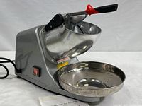 Full view of the silver electric ice crusher/shaver machine with power switch on side and attached stainless steel catch bowl.