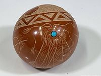 Oblique view of polished red clay ball with geometric border and inlaid turquoise bead