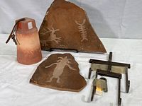 Display of two shale petroglyph stones, terra cotta bell, and brass display racks on white cloth background.
