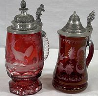 Two ruby glass beer steins with hinged metal lids, one with etched bird and nature scene and faceted base, the other with floral etched motifs and round base.