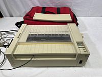 Front view of a vintage off-white Apple keyboard A9M0303 with a built-in printer and attached cables. Red carry bag and papers in background.